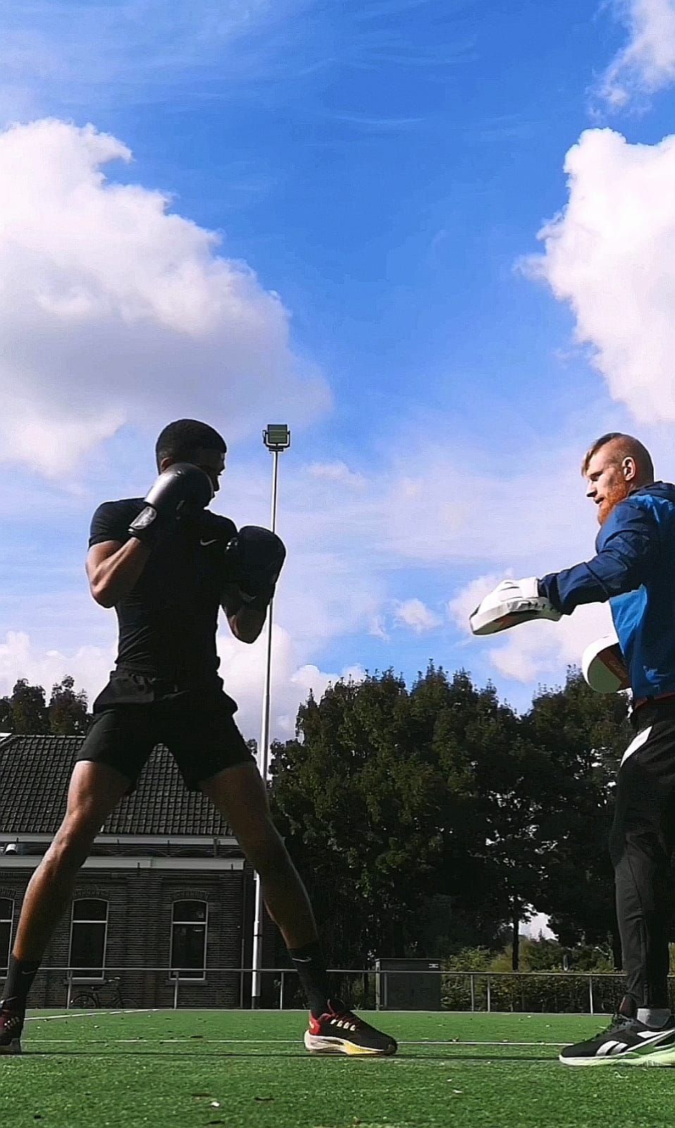 Manu boxeando con guantes frente a su entrenador, que le pone las manoplas, en un campo de césped bajo el cielo azul